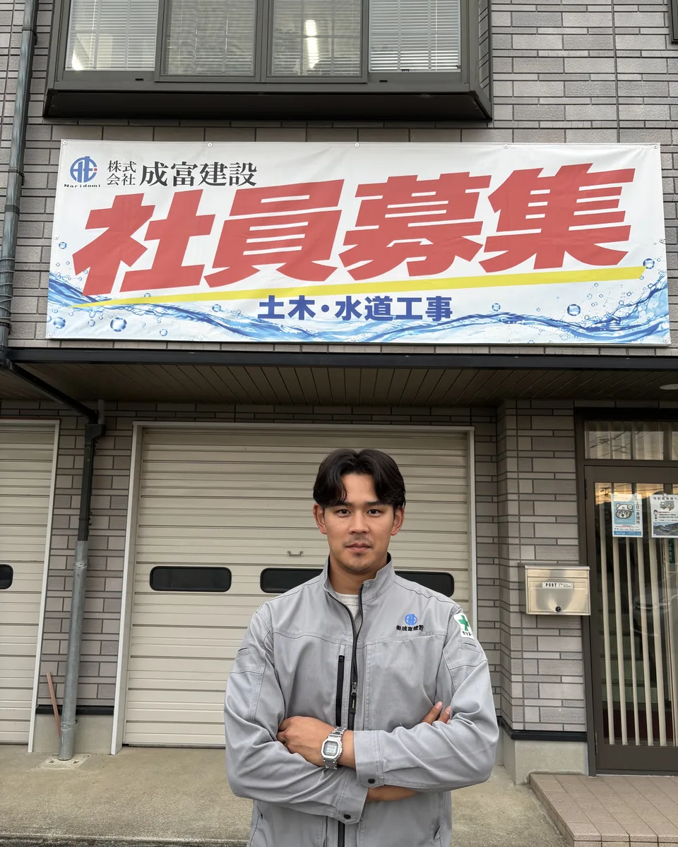 「社員募集」の横断幕の前で腕を組む成富建設の社員